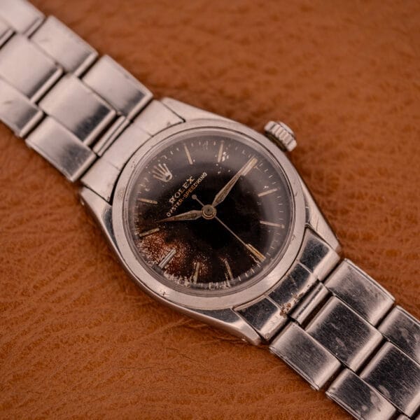 A Rolex Oyster Speedking Ref. 6420 Stainless Steel Black Dial wristwatch with a worn black dial is displayed on a brown textured surface.