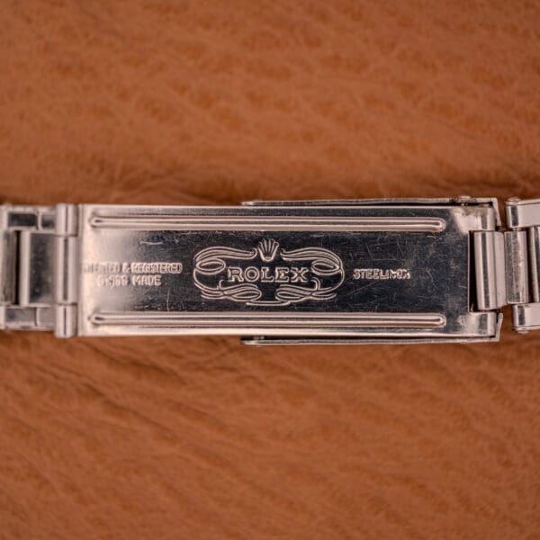 Close-up of the Rolex Oyster Speedking Ref. 6420 Stainless Steel Black Dial clasp, featuring the engraved Rolex logo, set against a brown textured background.