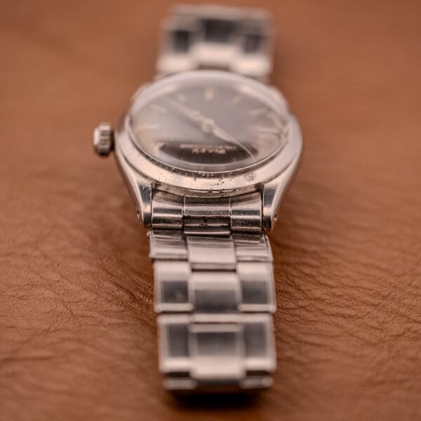 A Rolex Oyster Speedking Ref. 6420 Stainless Steel Black Dial wristwatch with a metal band is displayed elegantly on a brown leather surface.