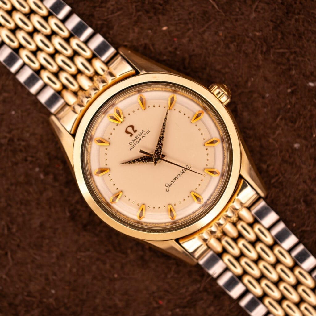 An Omega Seamaster "De Luxe" Ref. 2802 with Automatic Cal. 471 and a cream dial, featuring a two-tone metal band, is displayed elegantly on a brown surface.