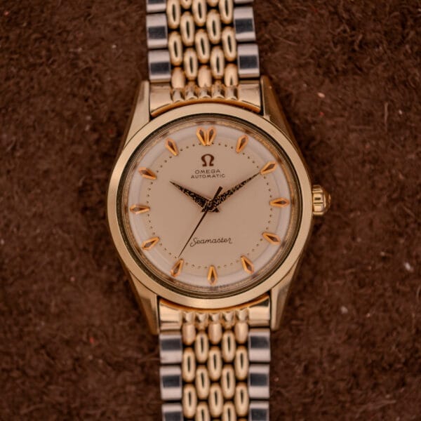 Omega Seamaster "De Luxe" Ref. 2802 watch with a metal band shown on a brown textured background at 10:09. Features Automatic Cal. 471 movement for precise timekeeping.