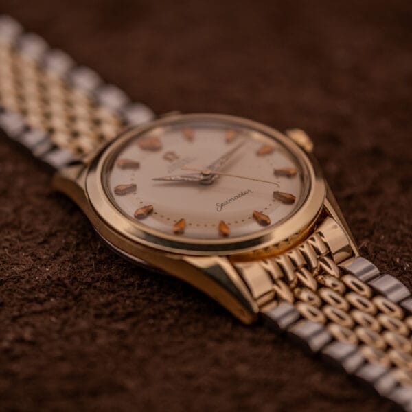 Omega Seamaster "De Luxe" Ref. 2802, a gold and silver wristwatch with a metal band, shown on a brown textured surface at 12:15. Features Automatic Cal. 471 movement for precise timekeeping.