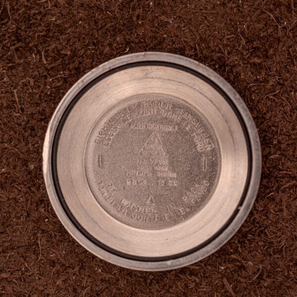 A metal geocaching container lid, echoing the robust design of an Omega Seamaster "De Luxe" Ref. 2802 Automatic Cal. 471, lies atop brown soil in an overhead view.