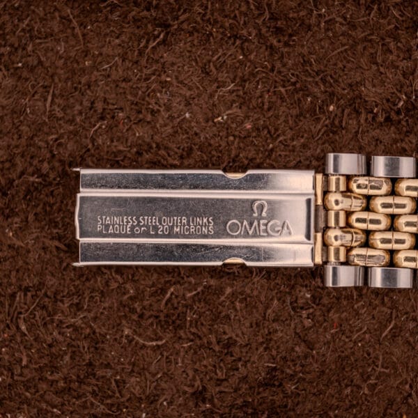 Close-up of an Omega Seamaster "De Luxe" Ref. 2802 Automatic Cal. 471 clasp, featuring engraved text and fine craftsmanship, set against a brown textured background.