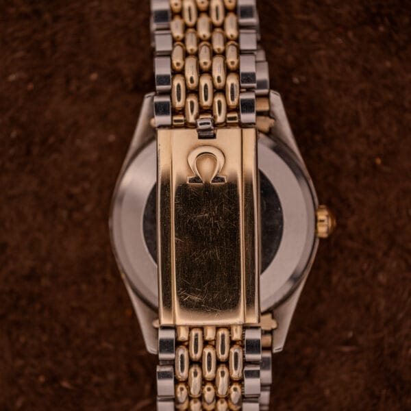 Close-up of the gold and silver clasp of an Omega Seamaster "De Luxe" Ref. 2802 Automatic Cal. 471 bracelet, engraved with the Omega logo, shown on a brown background.