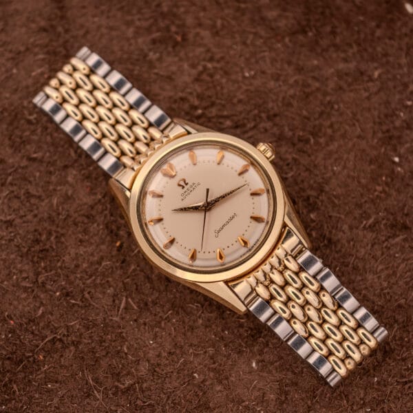 An Omega Seamaster "De Luxe" Ref. 2802 wristwatch with Automatic Cal. 471 movement and a metal bracelet, displayed on a brown textured surface.