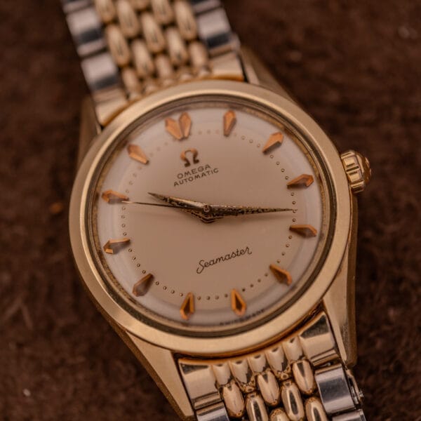 The Omega Seamaster "De Luxe" Ref. 2802 features a cream dial with gold indices, powered by Automatic Cal. 471, and is displayed on a brown surface.