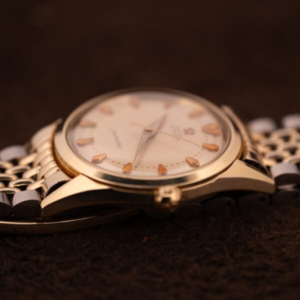 Close-up of an Omega Seamaster "De Luxe" Ref. 2802 Automatic Cal. 471 vintage gold wristwatch with a metal band, resting on a dark brown textured surface.