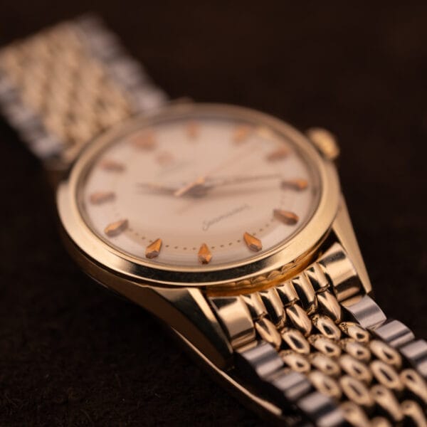 Close-up of an Omega Seamaster "De Luxe" Ref. 2802 wristwatch with a cream dial, gold hour markers, and Automatic Cal. 471 movement against a dark background.