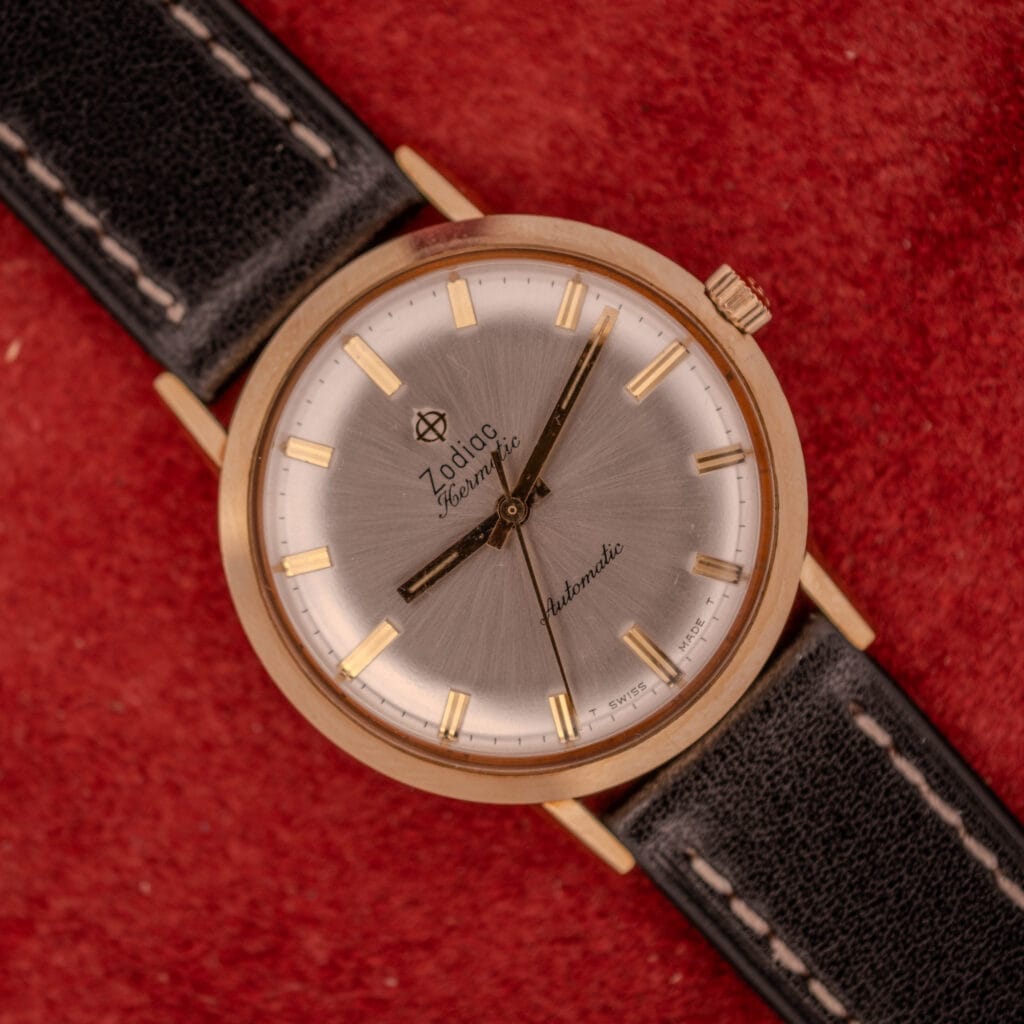 A Zodiac Hermetic Sunburst Dial Automatic wristwatch with a 10k gold filled case, silver face, and black leather strap displayed on a red background.