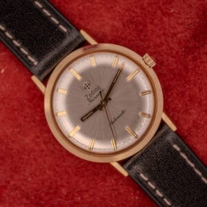A Zodiac Hermetic Sunburst Dial Automatic wristwatch with a 10k gold filled case, silver face, and black leather strap displayed on a red background.