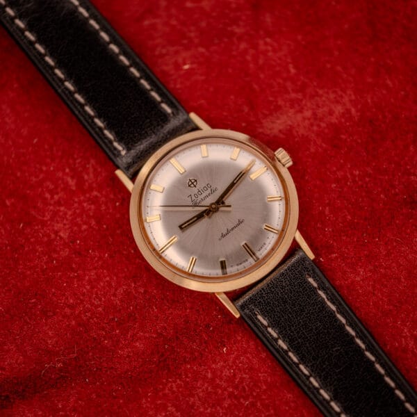 A Zodiac Hermetic Sunburst Dial 10k Gold Filled Case Automatic wristwatch with a black leather strap rests on a textured red surface.