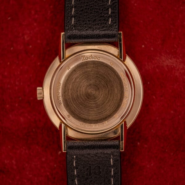 The Zodiac Hermetic Sunburst Dial 10k Gold Filled Case Automatic, featuring a black leather strap, is showcased on a red textured background.