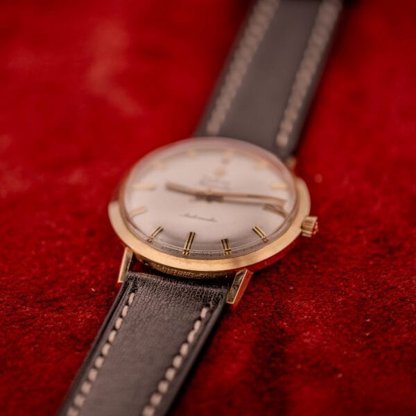 The Zodiac Hermetic Sunburst Dial automatic watch with a 10k gold filled case and black leather strap rests on a red textured surface.