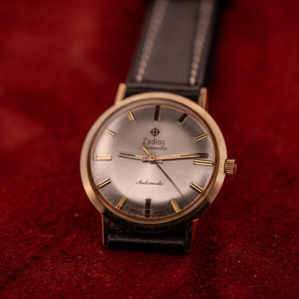 A Zodiac Hermetic Sunburst Dial 10k Gold Filled Case Automatic wristwatch with a black leather strap rests on a red surface.