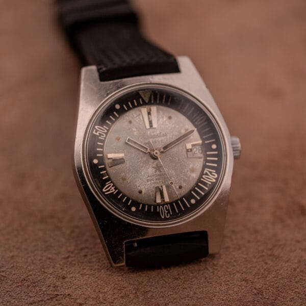The Aquastar 1701 Skin Diver 200 Meters Automatic, featuring a black tropic rubber strap and worn silver dial, is displayed on a brown textured surface.