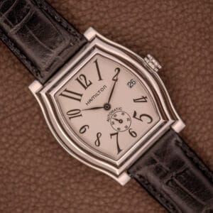 The Hamilton Dodson Automatic Stainless Steel Tonneau Case wristwatch, with a black leather strap and art deco-style dial, is displayed elegantly on a brown surface.