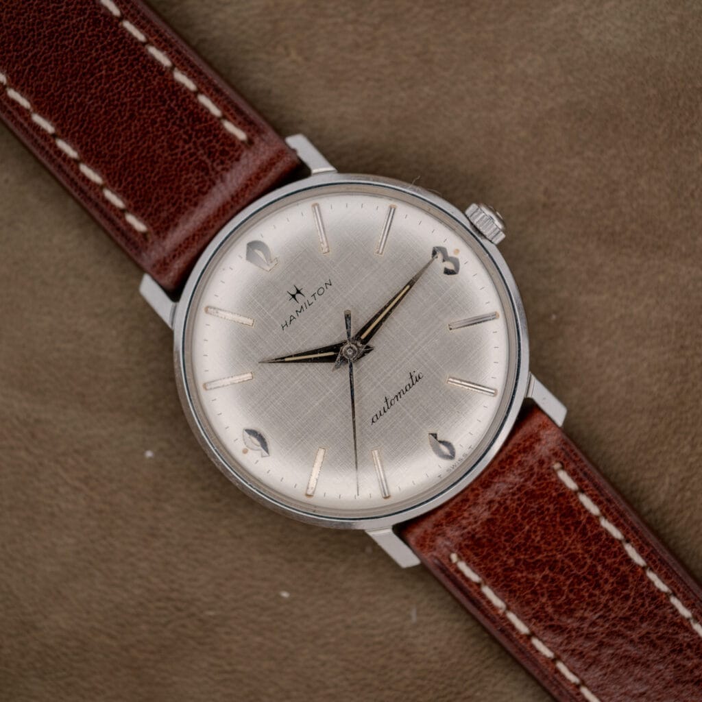 A Hamilton Automatic Silver Linen Dial Stainless Steel Cal. 689A wristwatch with a brown leather strap is displayed on a tan suede background.