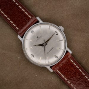 A Hamilton Automatic Silver Linen Dial Stainless Steel Cal. 689A wristwatch with a brown leather strap is displayed on a tan suede background.