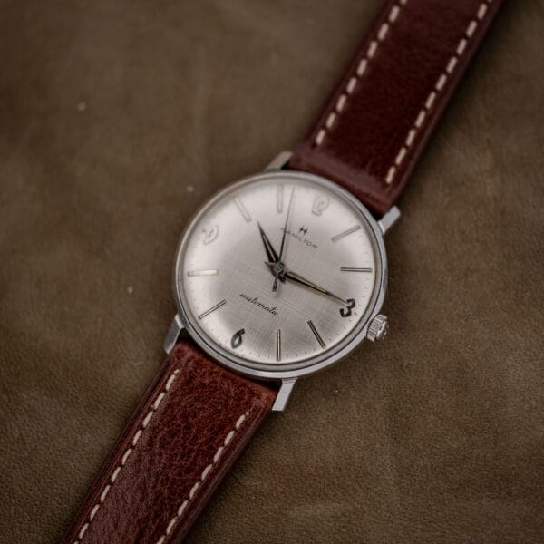 A Hamilton Automatic Silver Linen Dial Stainless Steel Cal. 689A wristwatch with a brown leather strap rests elegantly on a suede surface.