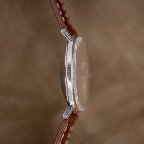 Side view of the Hamilton Automatic Silver Linen Dial Stainless Steel Cal. 689A, showcasing its slim silver case and brown leather strap against a brown background.