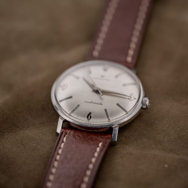 The Hamilton Automatic Silver Linen Dial Stainless Steel Cal. 689A wristwatch, paired with a brown leather strap, rests elegantly on a brown fabric surface.