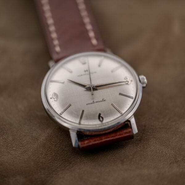 The Hamilton Automatic Silver Linen Dial Stainless Steel Cal. 689A watch rests elegantly on a brown suede surface.