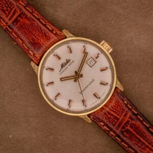The Mido Ocean Star Powerwind Date Ladies Gold Tone wristwatch with a brown leather strap is elegantly displayed on a brown surface.