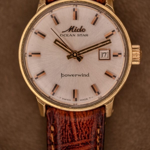 The Mido Ocean Star Powerwind Date Ladies Gold Tone watch pairs a brown leather strap with a date window, all set against a rich brown background.
