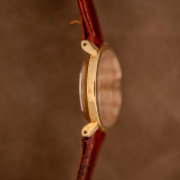 Side view of the Mido Ocean Star Powerwind Date Ladies Gold Tone wristwatch featuring a brown leather strap, inspired by classic elegance, set against a blurred brown background.