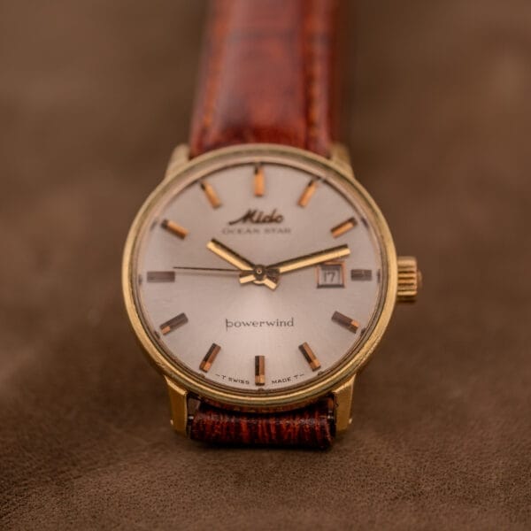 A Mido Ocean Star Powerwind Date Ladies Gold Tone watch with a brown leather strap rests elegantly on a brown surface.