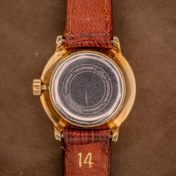 Back of a vintage Mido Ocean Star Powerwind Date Ladies Gold Tone wristwatch with a brown leather strap marked 14, highlighting engraved details on the gold-tone case.