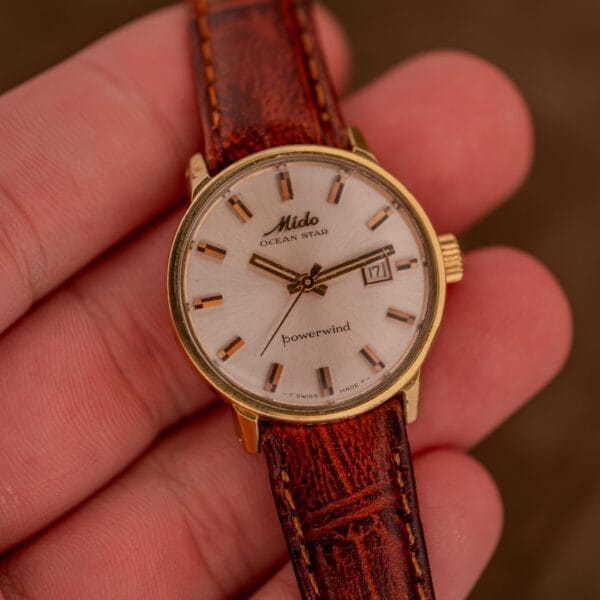 A hand holds a Mido Ocean Star Powerwind Date Ladies Gold Tone watch with a brown leather strap.