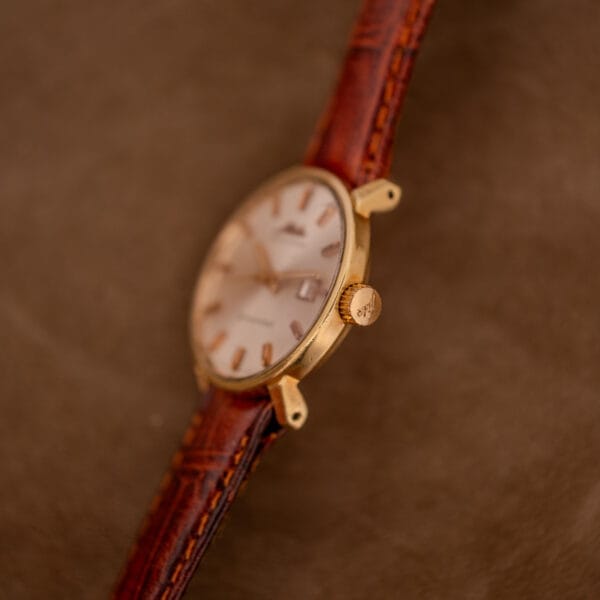 A Mido Ocean Star Powerwind Date Ladies Gold Tone wristwatch with a brown leather strap is elegantly displayed on a brown suede background.