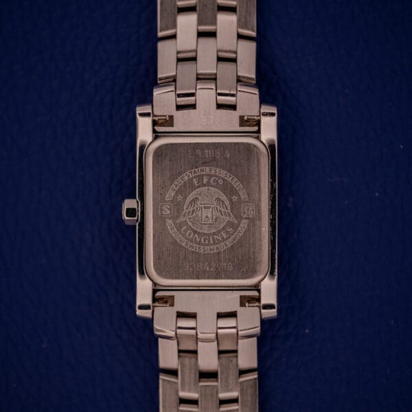 A close-up of the back of a Longines Dolce Vita L5.155.4 stainless steel tank case wristwatch, set on a dark blue textured surface.