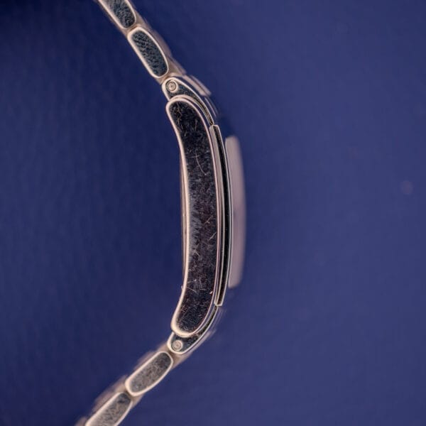 A close-up of a scratched stainless steel clasp, likely from a Longines Dolce Vita L5.155.4 with its signature tank case, set against a textured dark blue background.