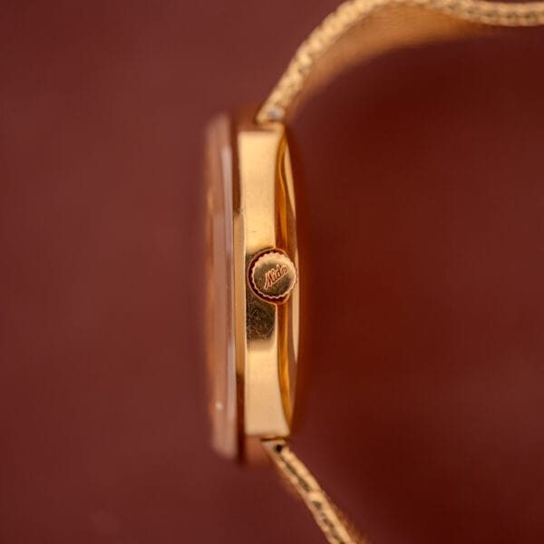 Side view of the Mido Commander Automatic Datoday Ref. 8429 with textured crown and elegant Milanese strap, set against a brown background.