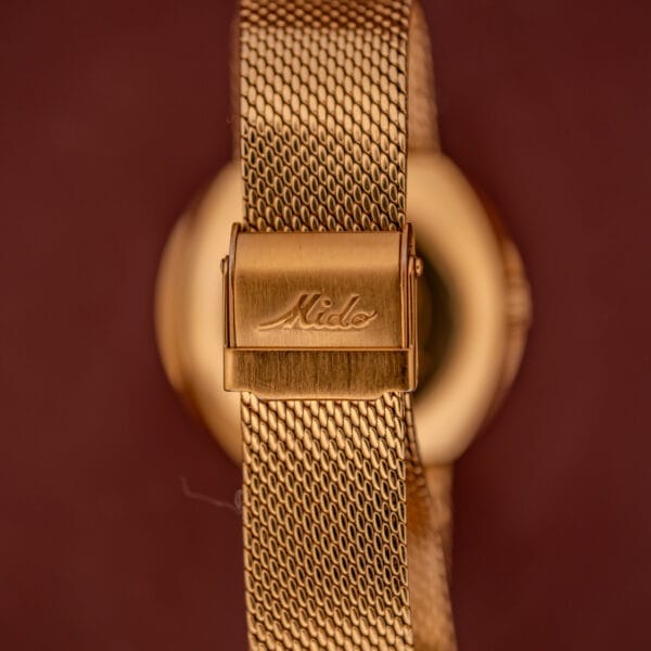 Close-up of the Mido Commander Automatic Datoday Ref. 8429 with a gold Milanese strap and "Mido" engraved on the clasp, set against a brown background, showcases the watch's elegance.
