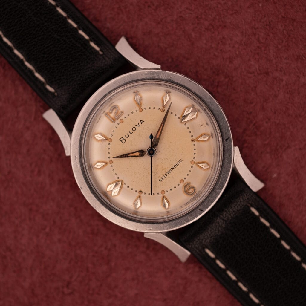 The Bulova Self-Winding Cal. 11AFAC features a stainless steel case with fancy lugs, a beige dial, and a black leather strap, displayed on a maroon background.