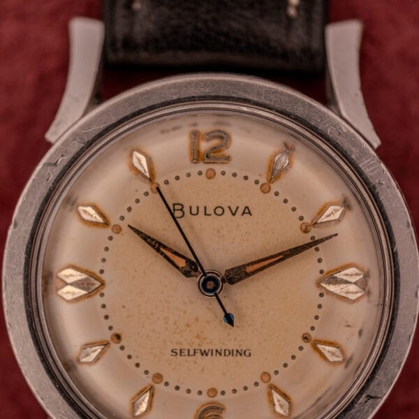 Close-up of a Bulova Self-Winding Cal. 11AFAC watch with gold markers, fancy lugs, and a stainless steel case, set against a red background.