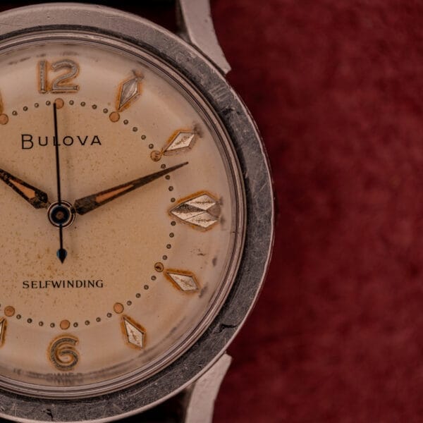 Close-up of a Bulova Self-Winding Cal. 11AFAC watch featuring fancy lugs, gold details, and a polished stainless steel case, set against a maroon textured background.