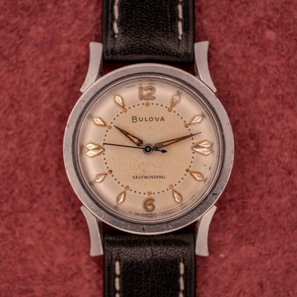 A Bulova Self-Winding Cal. 11AFAC wristwatch with fancy lugs, cream dial, black leather strap, and stainless steel case rests on a textured red surface.