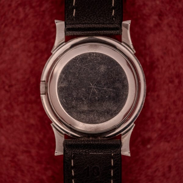 The scratched back of a Bulova Self-Winding Cal. 11AFAC with stainless steel case and fancy lugs rests on a red textured background, paired with a black leather strap.