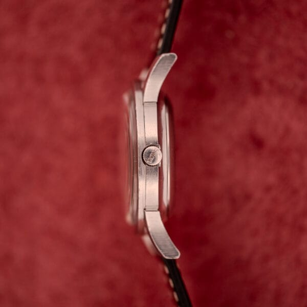 Side view of the Bulova Self-Winding Cal. 11AFAC wristwatch with a stainless steel case and fancy lugs, displayed on a black strap against a textured red background.