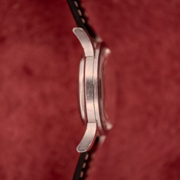 Side view of a Bulova Self-Winding Cal. 11AFAC wristwatch with a stainless steel case and fancy lugs, displayed against a red textured background.