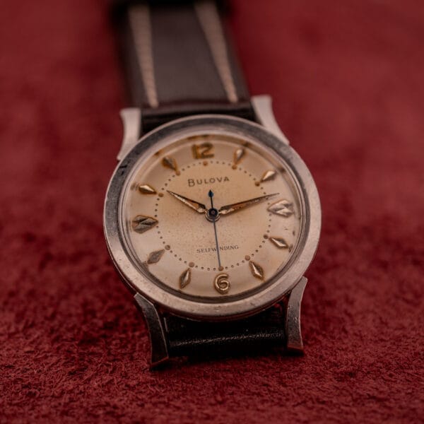 Bulova Self-Winding Cal. 11AFAC wristwatch with a stainless steel case and fancy lugs, shown on a red textured surface.