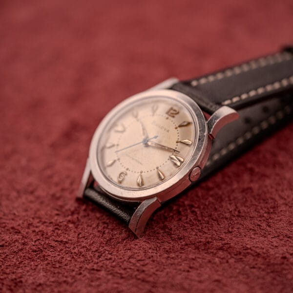 Bulova Self-Winding Cal. 11AFAC wristwatch with fancy lugs, cream dial, and black leather strap in a stainless steel case, displayed on a textured red surface.