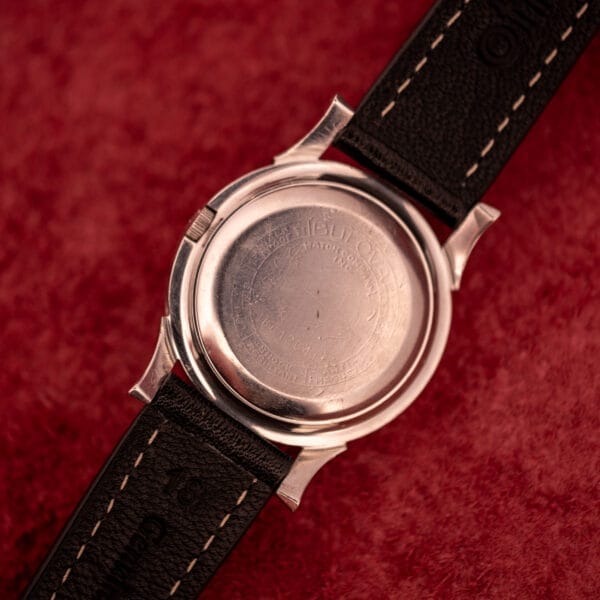 A close-up of a Bulova Self-Winding Cal. 11AFAC wristwatch with black leather straps and a stainless steel case with fancy lugs, resting on a textured red surface.