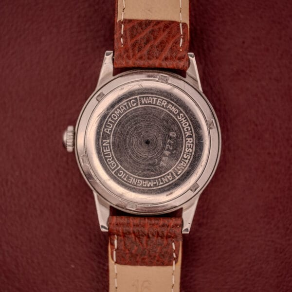 Back of a Gruen Precision Auto-Wind Copper Dial Stainless Steel Cal. 400 wristwatch with brown leather strap on a red surface, showing engraved text about its automatic movement and stainless steel build.