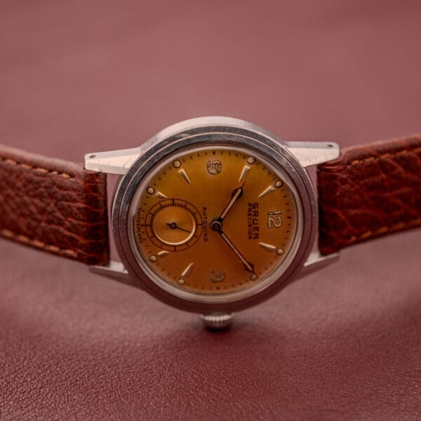 A Gruen Precision Auto-Wind Copper Dial Stainless Steel Cal. 400 wristwatch with a brown leather strap sits elegantly on a reddish surface.